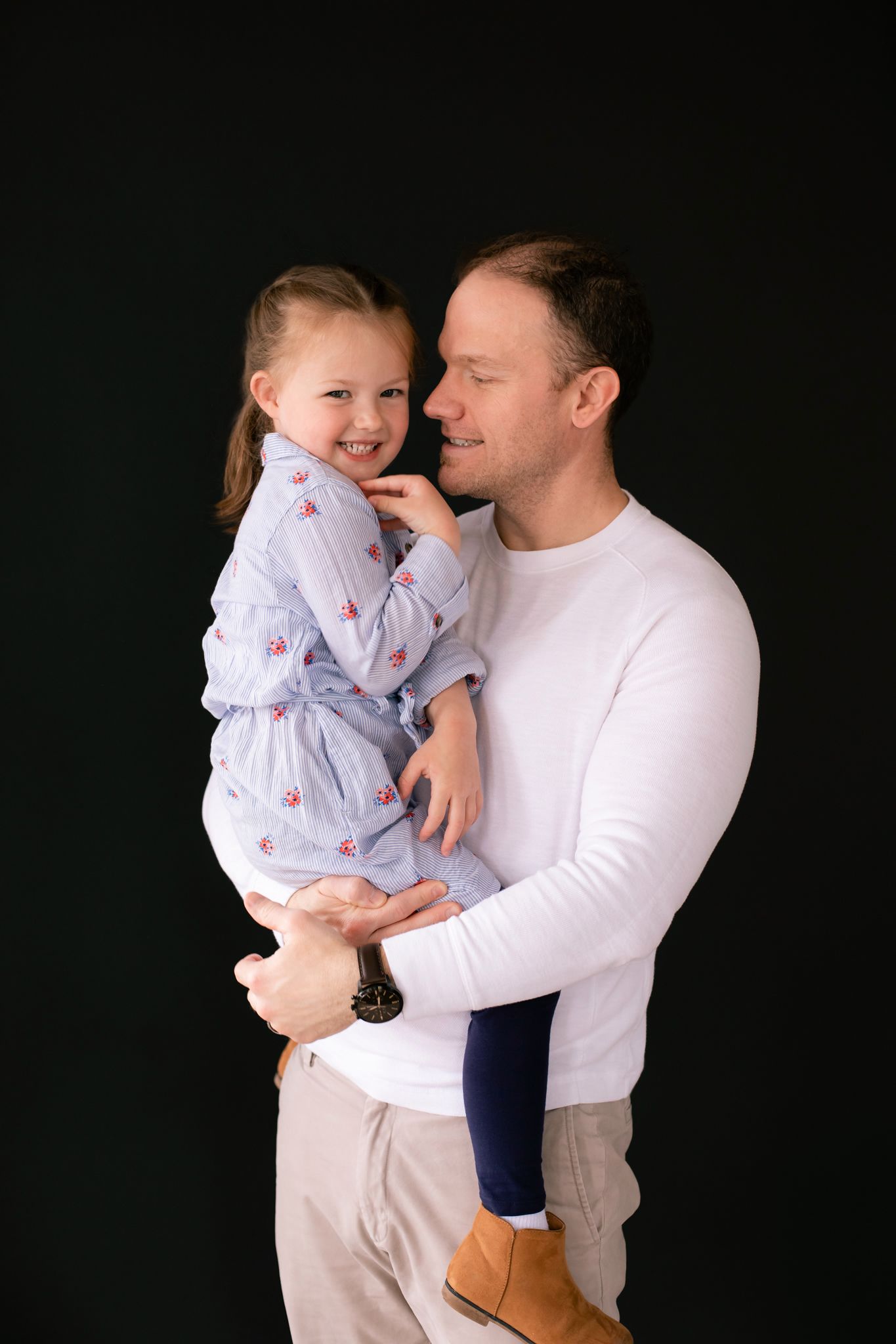 Loving father holding smiling young daughter in floral dress against black background portrait.