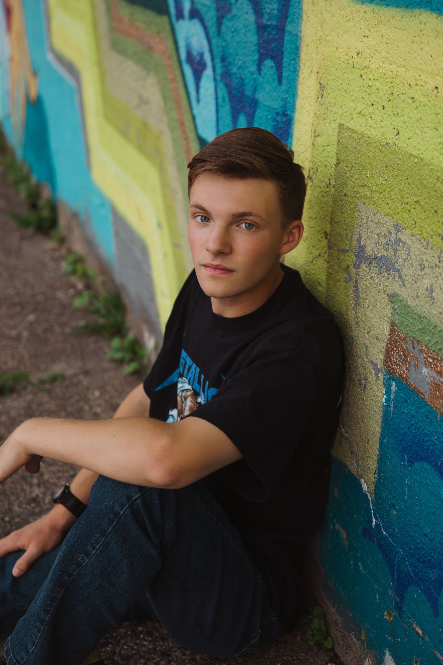 Teen boy in black t-shirt sits against colorful graffiti wall outdoors, looking at camera.