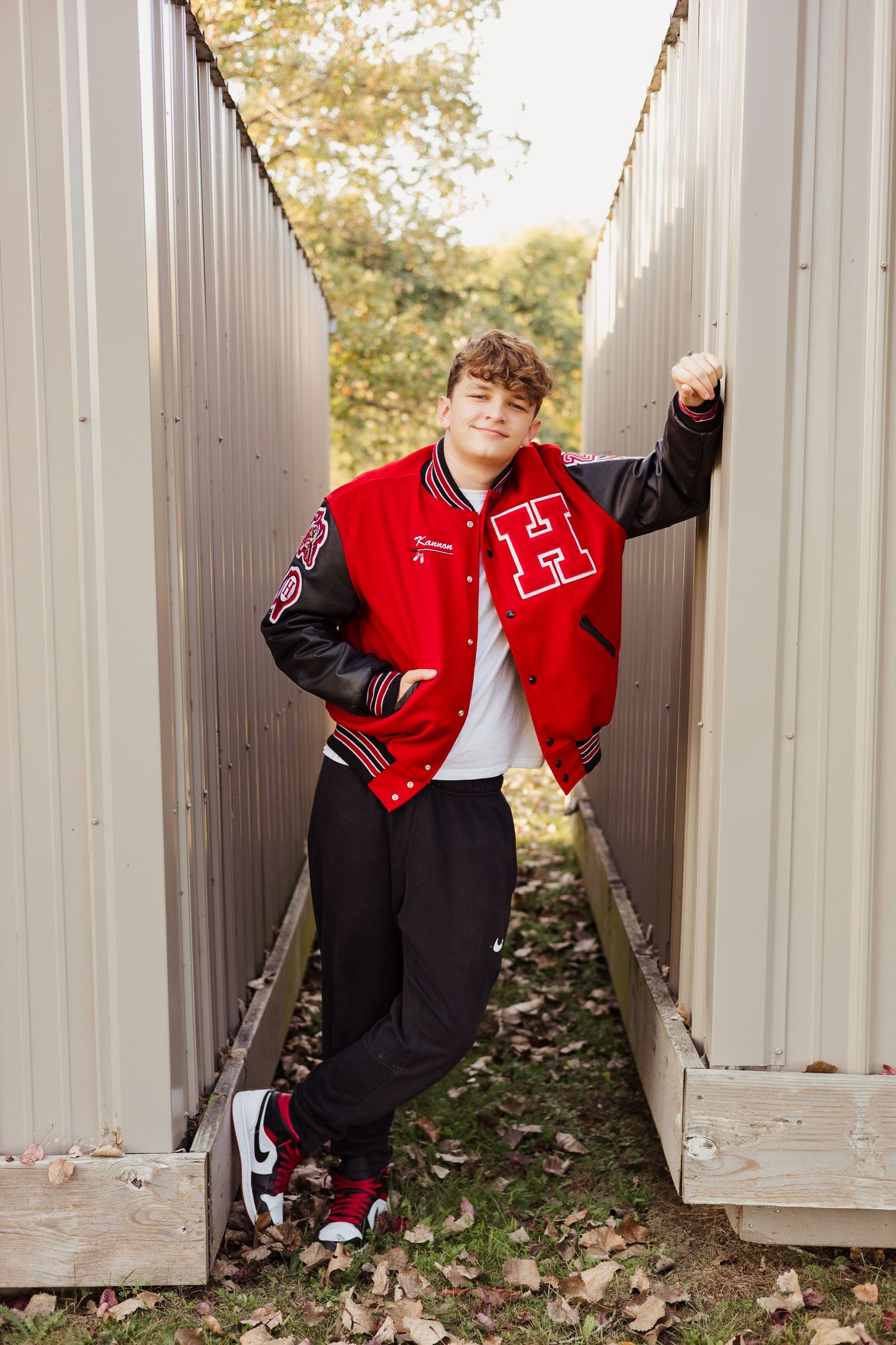 Teen boy in red varsity jacket with H letter poses between metal sheds surrounded by autumn leaves.