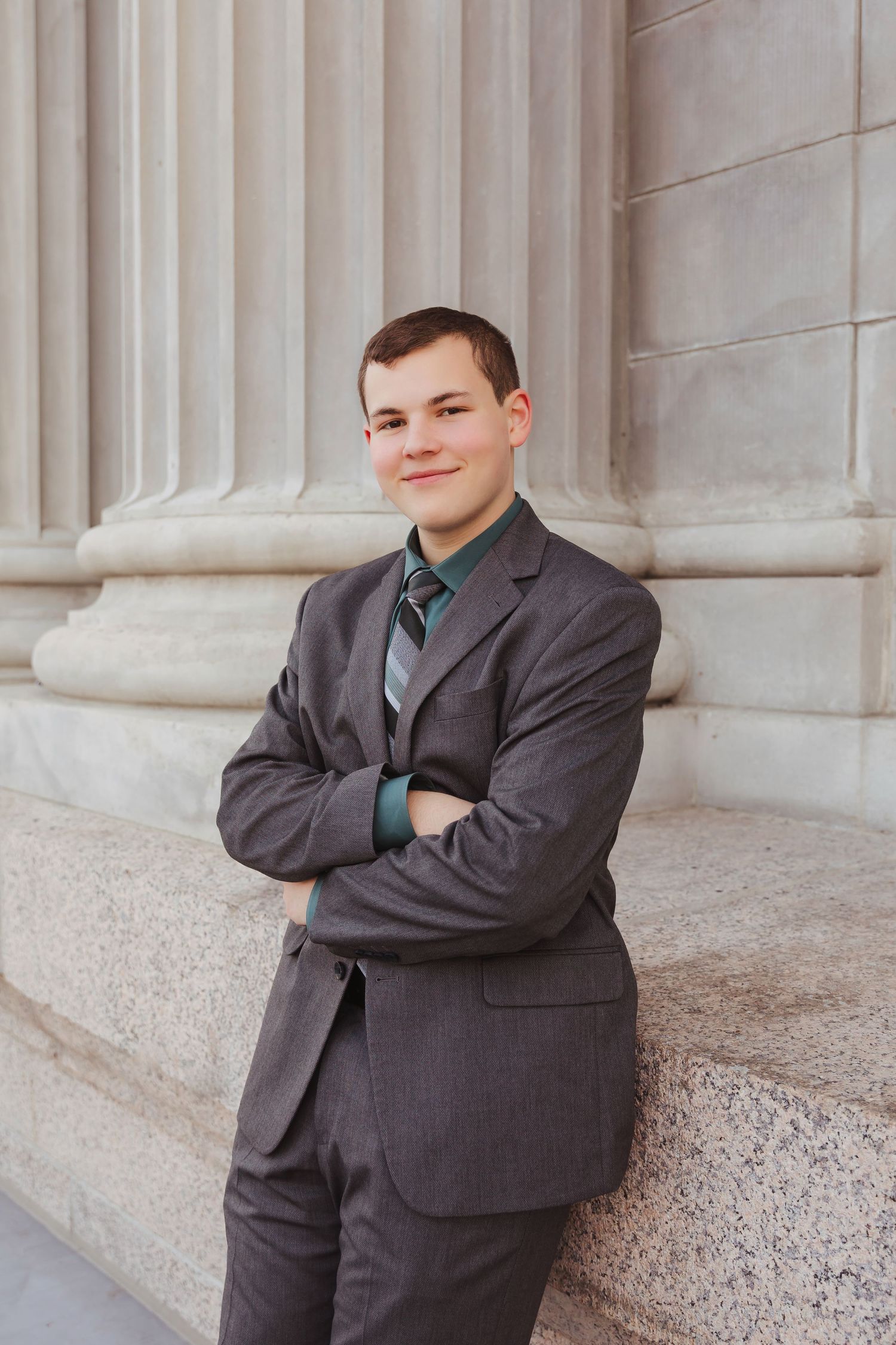 Young man in gray suit and teal tie stands confidently with arms crossed beside stone columns.