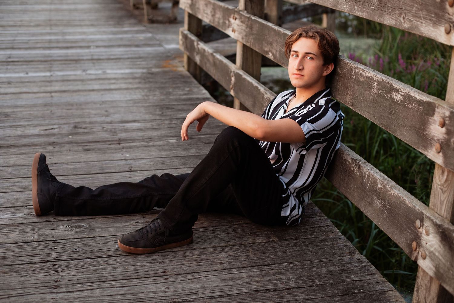 A teenage boy in a striped shirt and black pants sits relaxed on a wooden boardwalk, leaning against a fence.