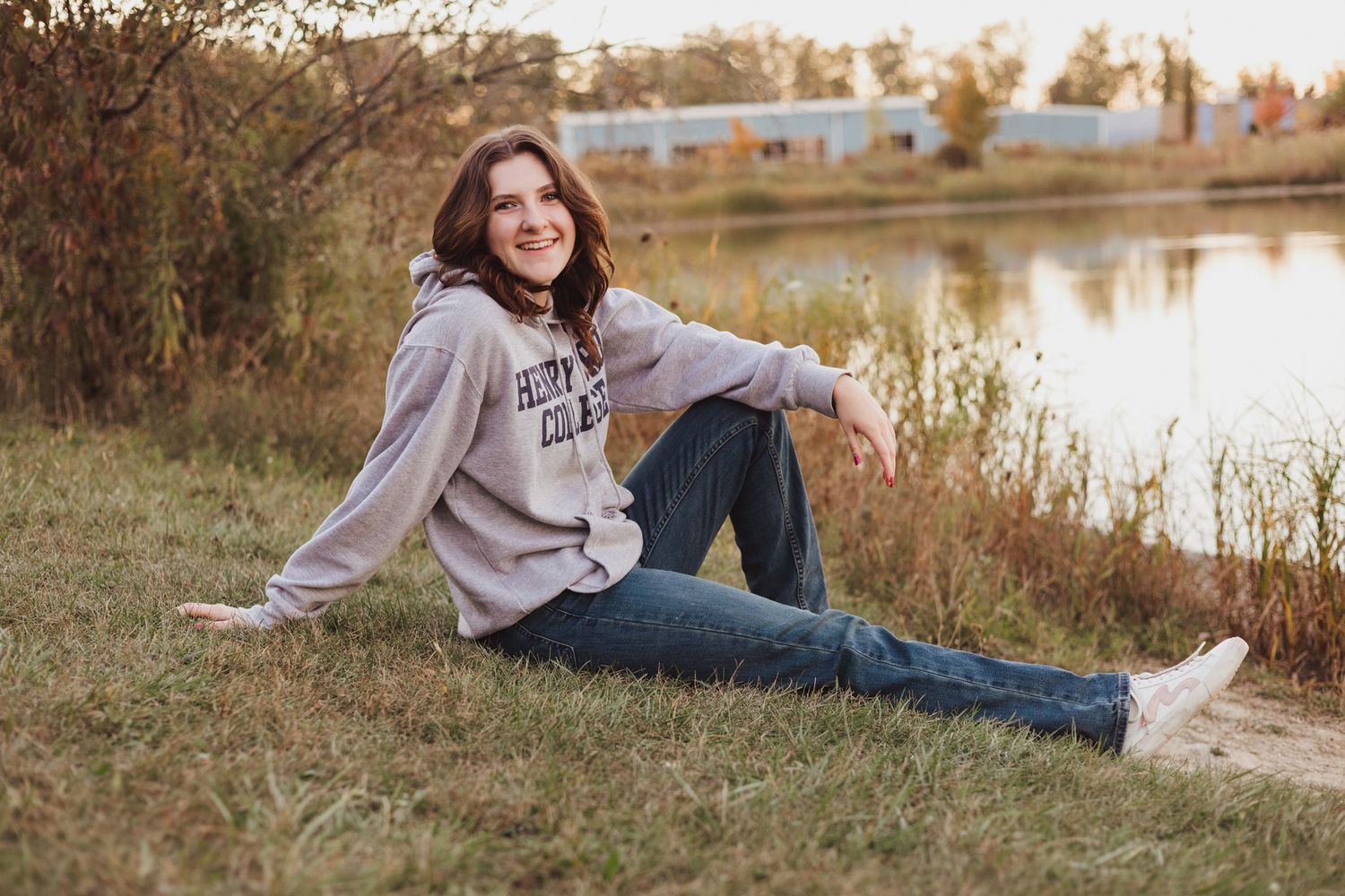 Young woman in gray hoodie and jeans sitting on grass by a lake, smiling outdoors in autumn.
