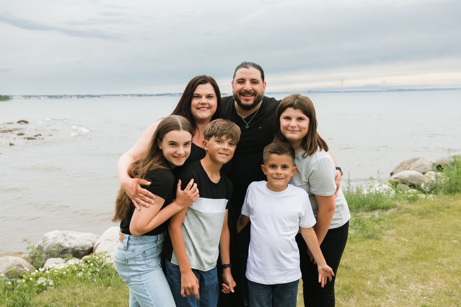 Family of five smiling together near a lakeside, with cloudy skies and rocky shore in the background.