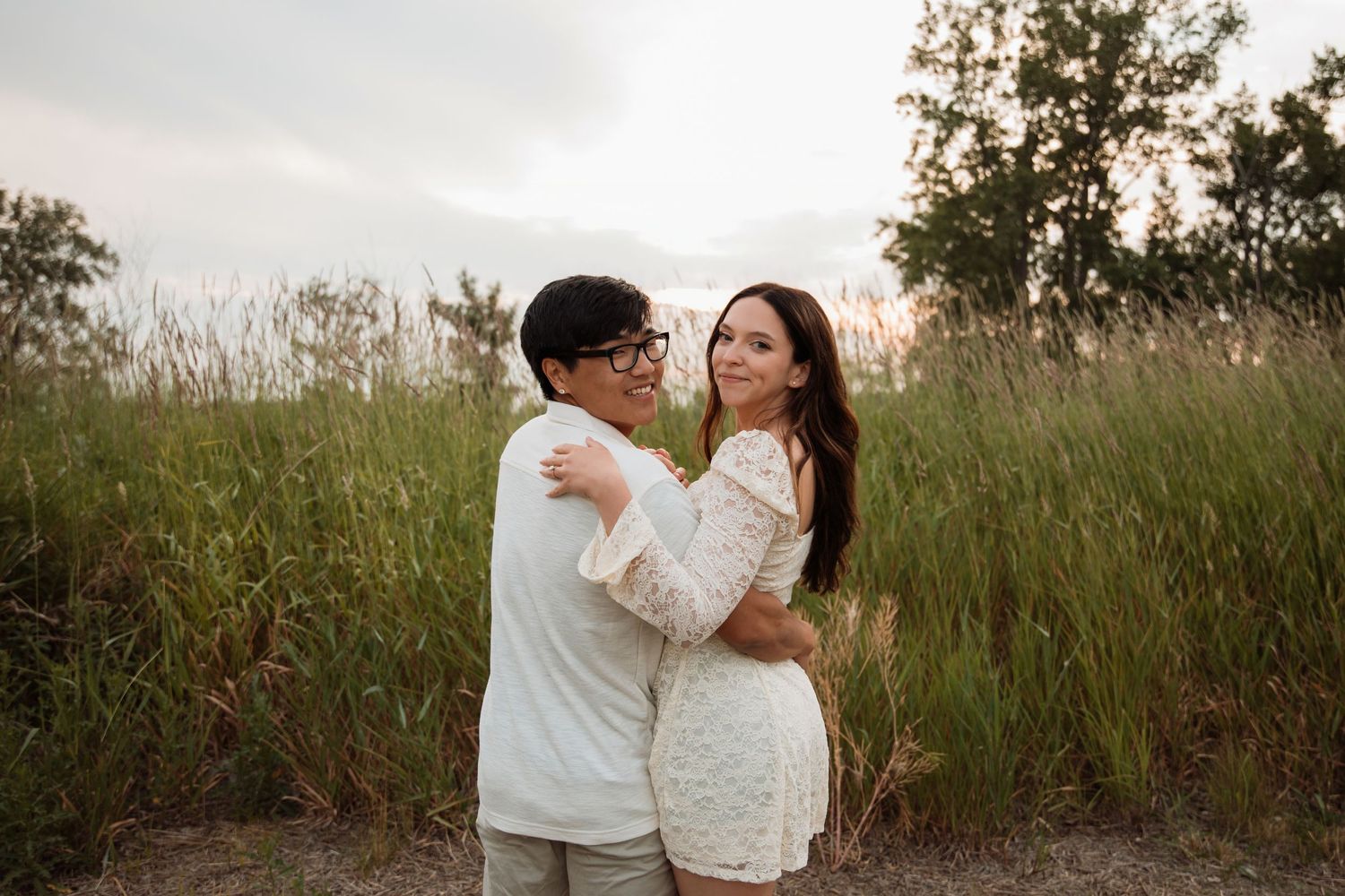 Couple embracing and smiling in tall green grass field at sunset, woman wearing white lace dress.