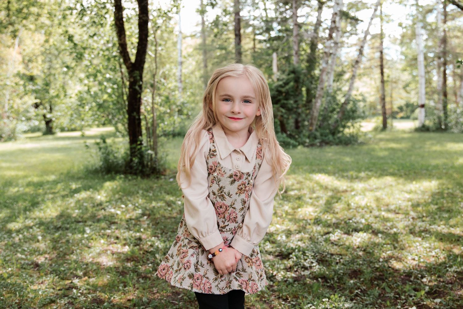 Young blonde girl in floral dress and cream top smiling outdoors in a sunny green park setting.