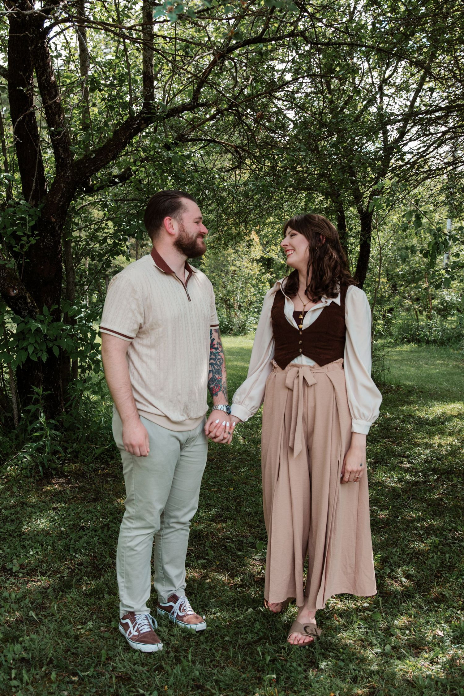 A couple holds hands and smiles at each other in a lush green garden, dressed in casual and folk-inspired outfits.