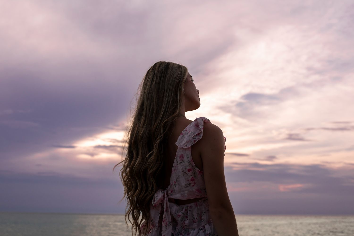 Young woman with long wavy hair in floral dress gazing at a purple sunset over the ocean.