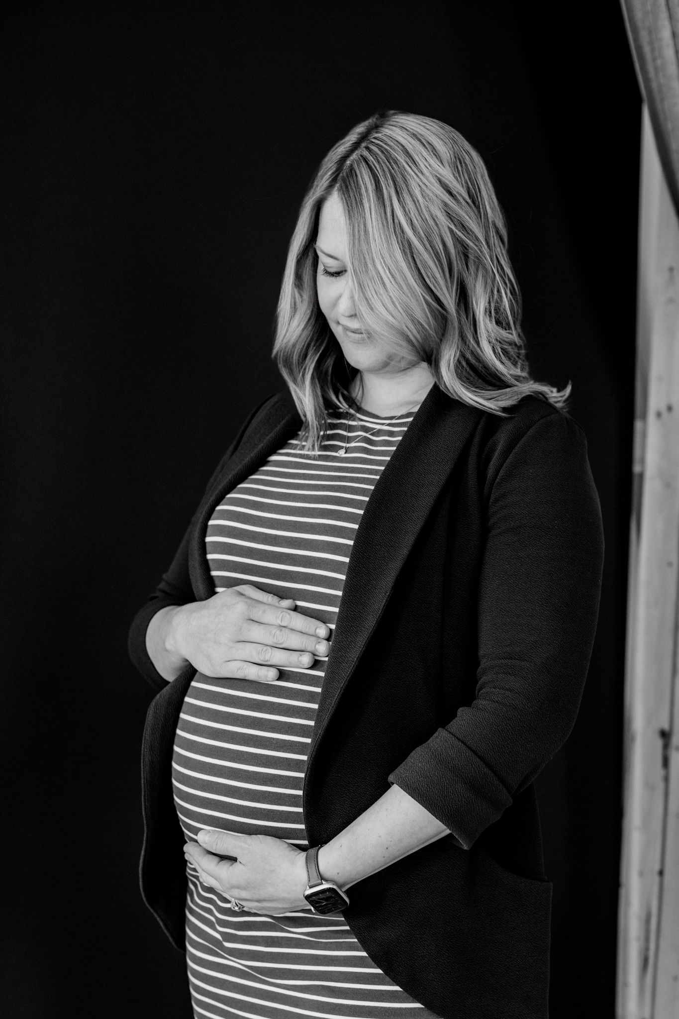 Pregnant woman in striped dress and black blazer tenderly holding baby bump in black and white portrait.