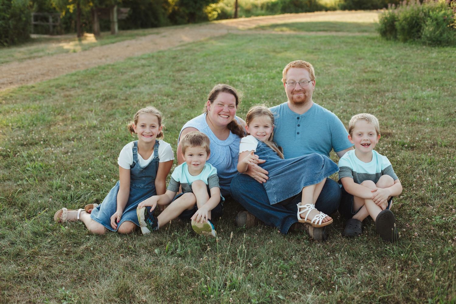 Happy family of six poses outdoors on green grass, parents with four young children smiling together in casual coordinated outfits.