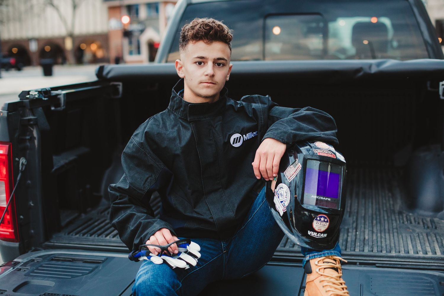 Young welder sitting in truck bed holding welding helmet and gloves wearing Miller Electric jacket outdoors.