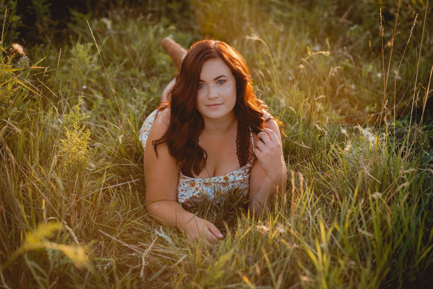 Red-haired woman in floral sundress lying in tall golden grass during warm summer sunset.