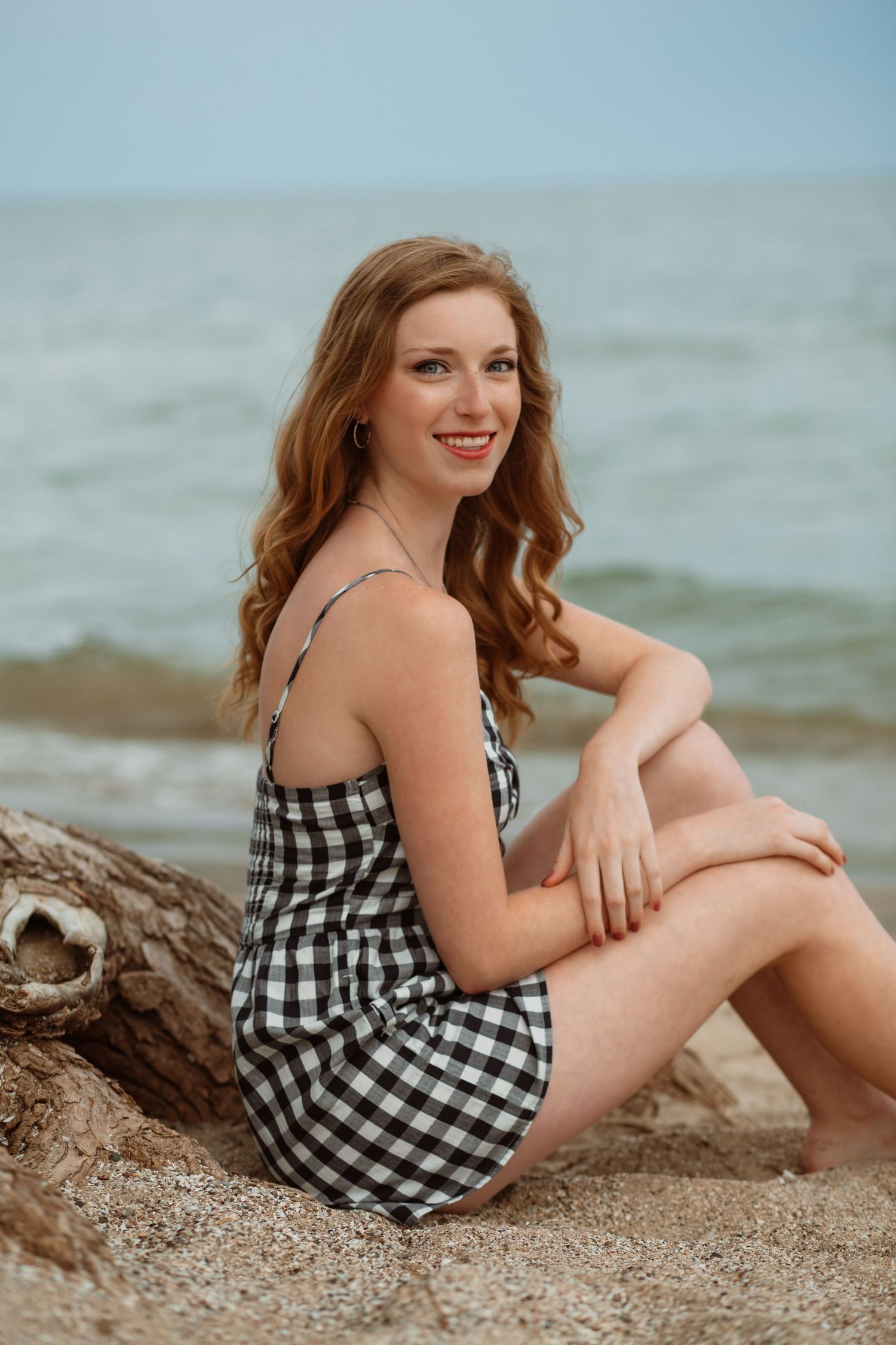 Young woman in black gingham dress sitting on driftwood log on sandy beach by ocean.