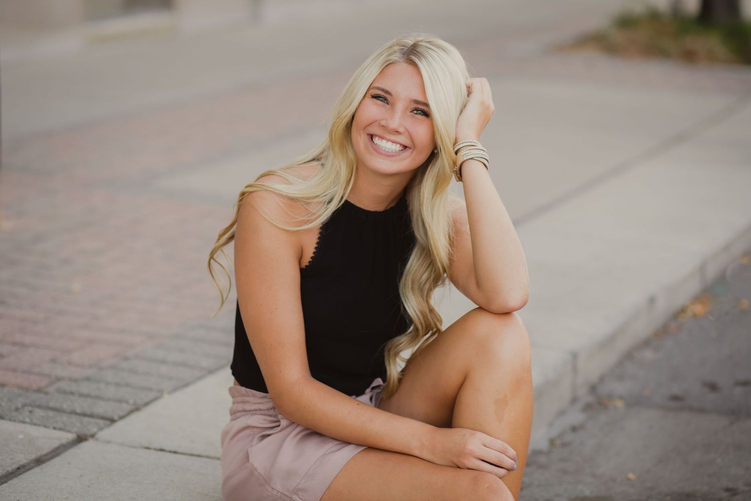 Blonde woman in black top and pink skirt sits smiling on urban sidewalk.