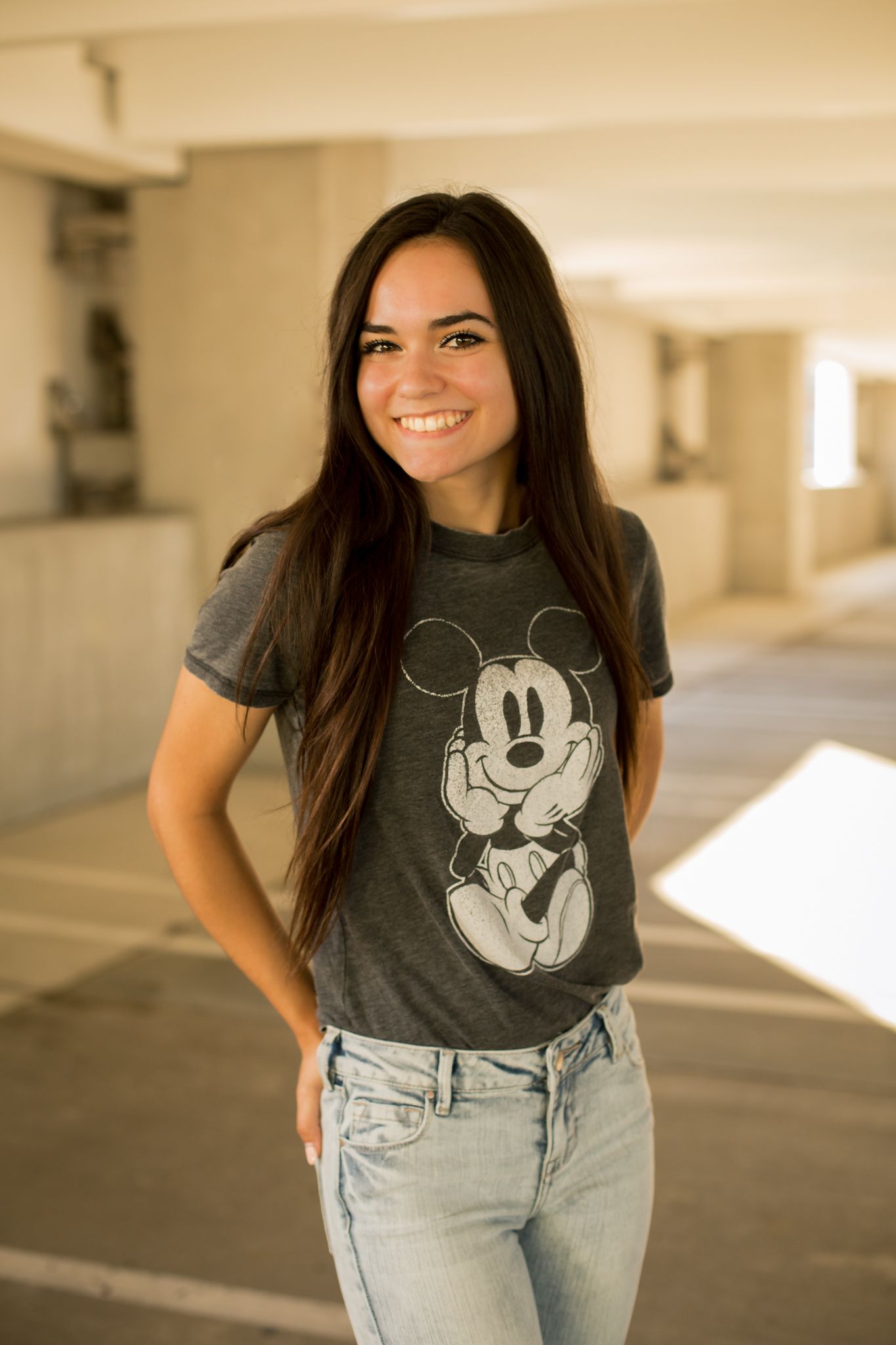 Smiling brunette woman in Mickey Mouse graphic tee and jeans stands in sunny parking garage.