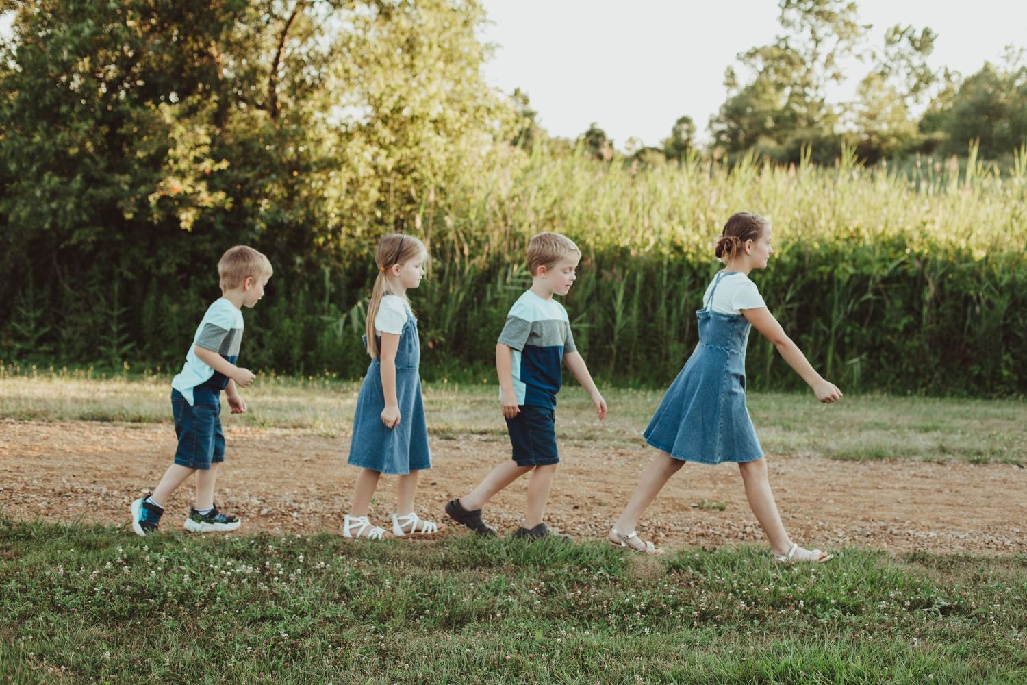 Four children in matching outfits walk in a line across a grassy field during a sunny outdoor family photo session.