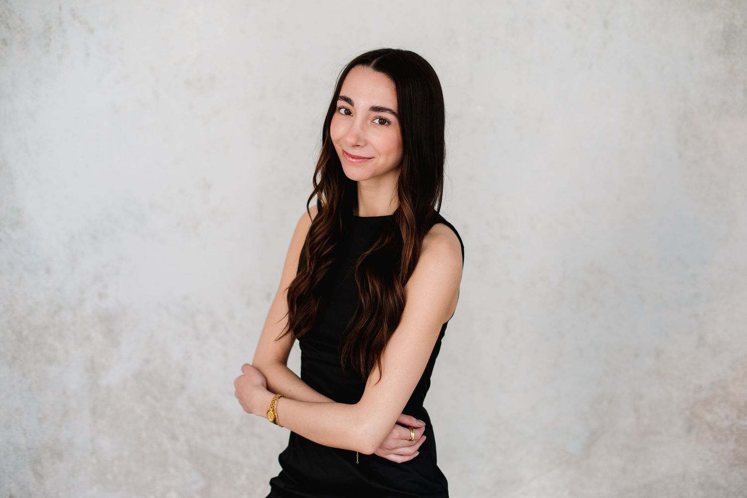A young woman with long dark wavy hair crosses her arms and smiles while posing against a light gray textured wall.