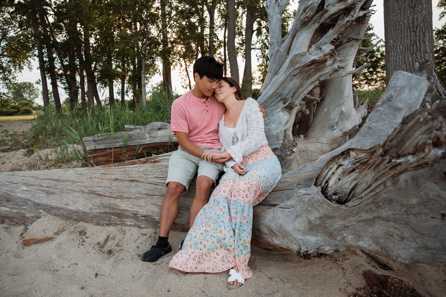 Couple sitting together on driftwood log on sandy beach surrounded by trees.