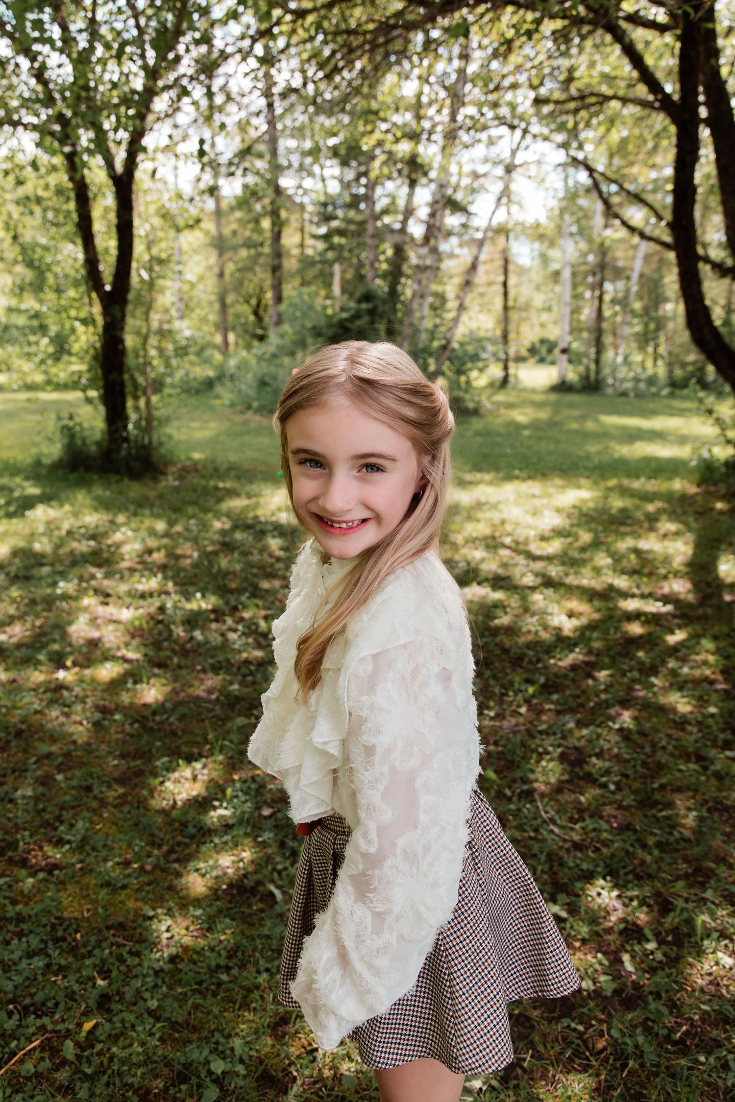 Smiling young girl with blonde hair wearing a white ruffled top and plaid skirt standing in a sunny green park.