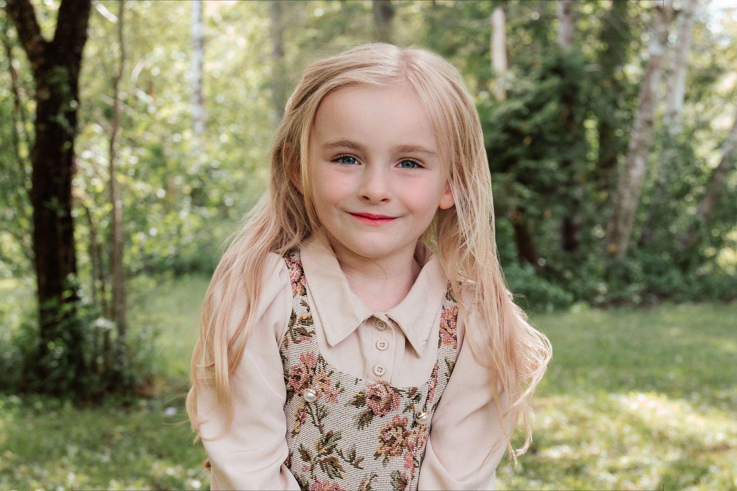 Young blonde girl smiling outdoors in floral dress over cream blouse, green trees in background.
