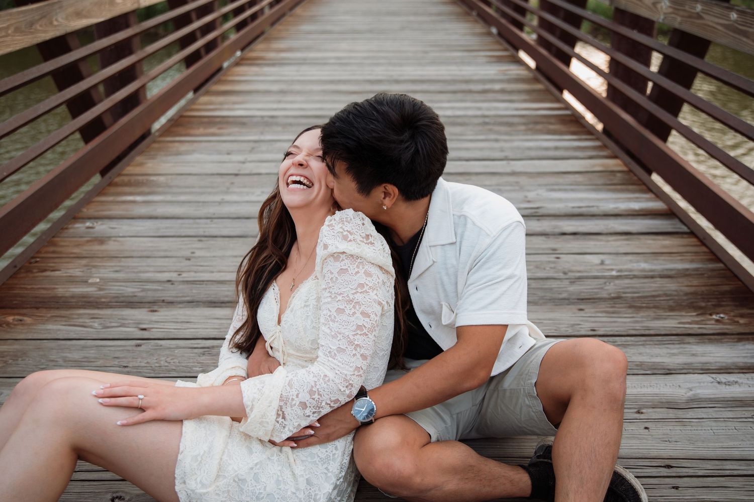 Couple sitting on wooden bridge, laughing and embracing, woman in white lace dress, man in light blue shirt and gray shorts.