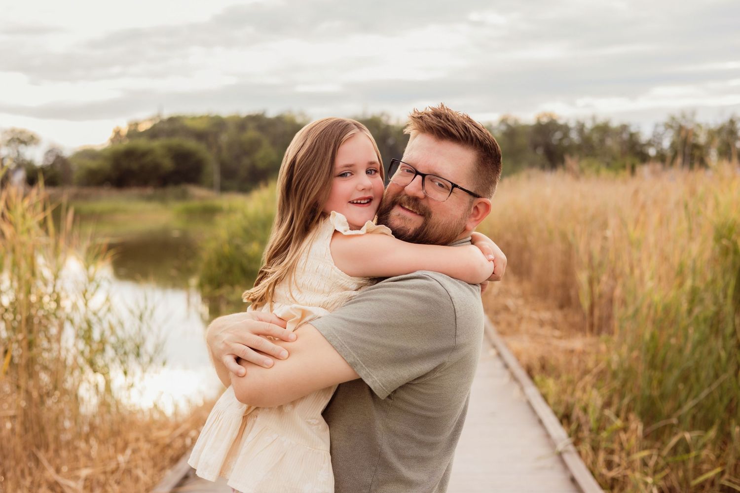 A father hugs his smiling daughter on a wooden boardwalk surrounded by golden grass near a pond.