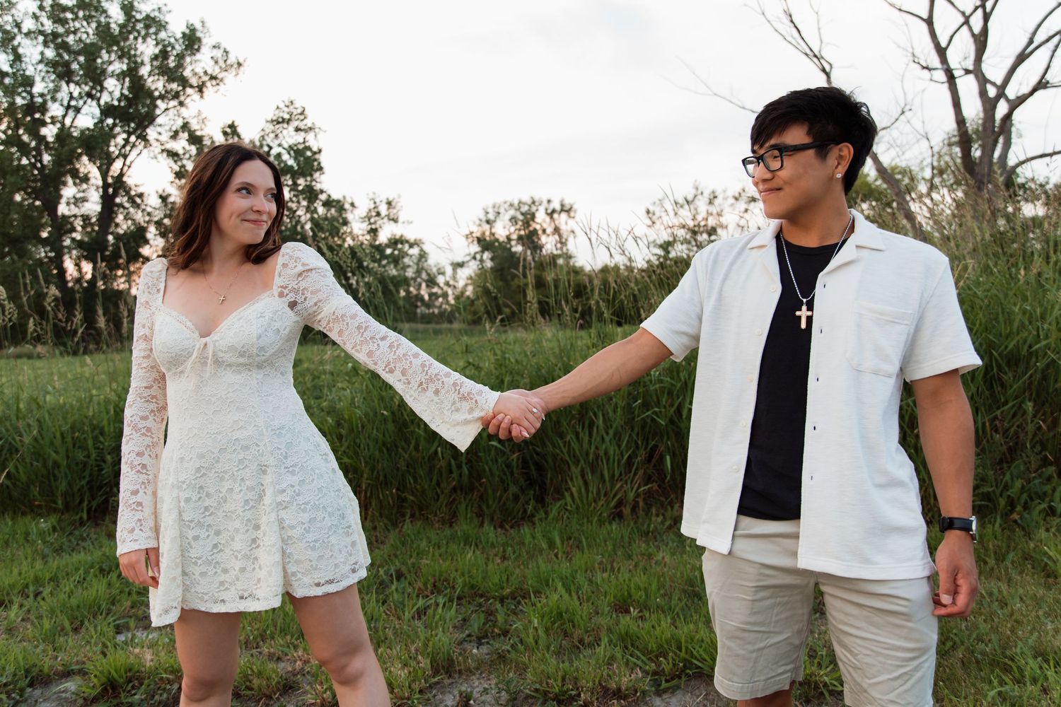 Couple holding hands and smiling at each other in a lush green outdoor setting at dusk.