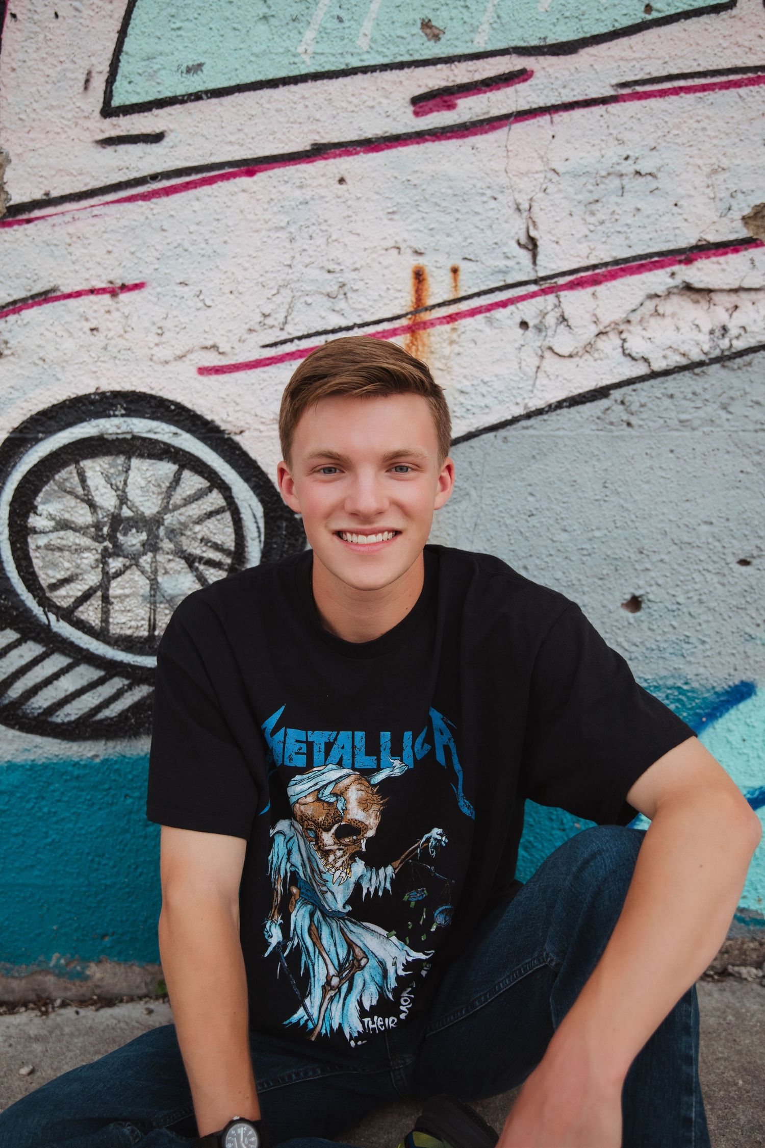 Smiling teen in Metallica t-shirt sits against colorful graffiti wall outdoors.