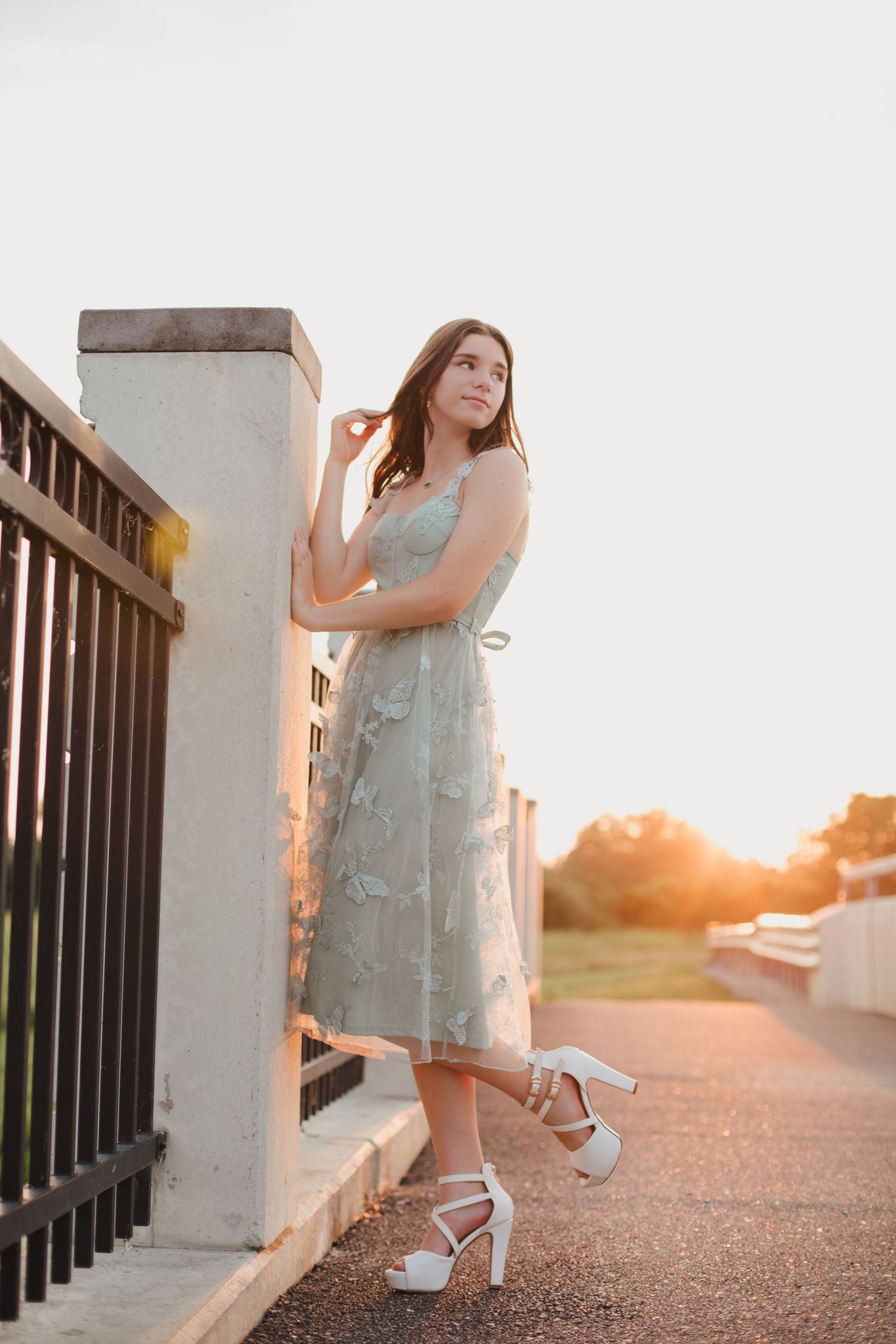 Young woman in mint green floral dress and white heels poses by iron fence at golden hour sunset.
