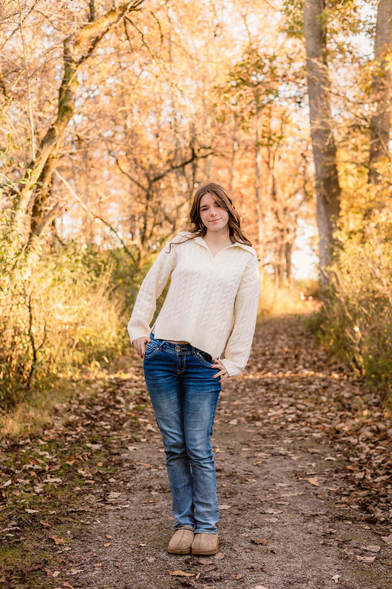 Young woman in cream sweater and jeans standing on a forest path surrounded by golden autumn foliage.