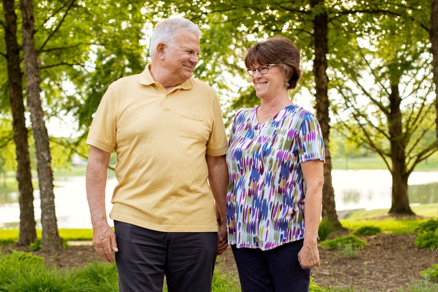 Elderly couple smiling and holding hands while walking together in a lush green park on a sunny day.