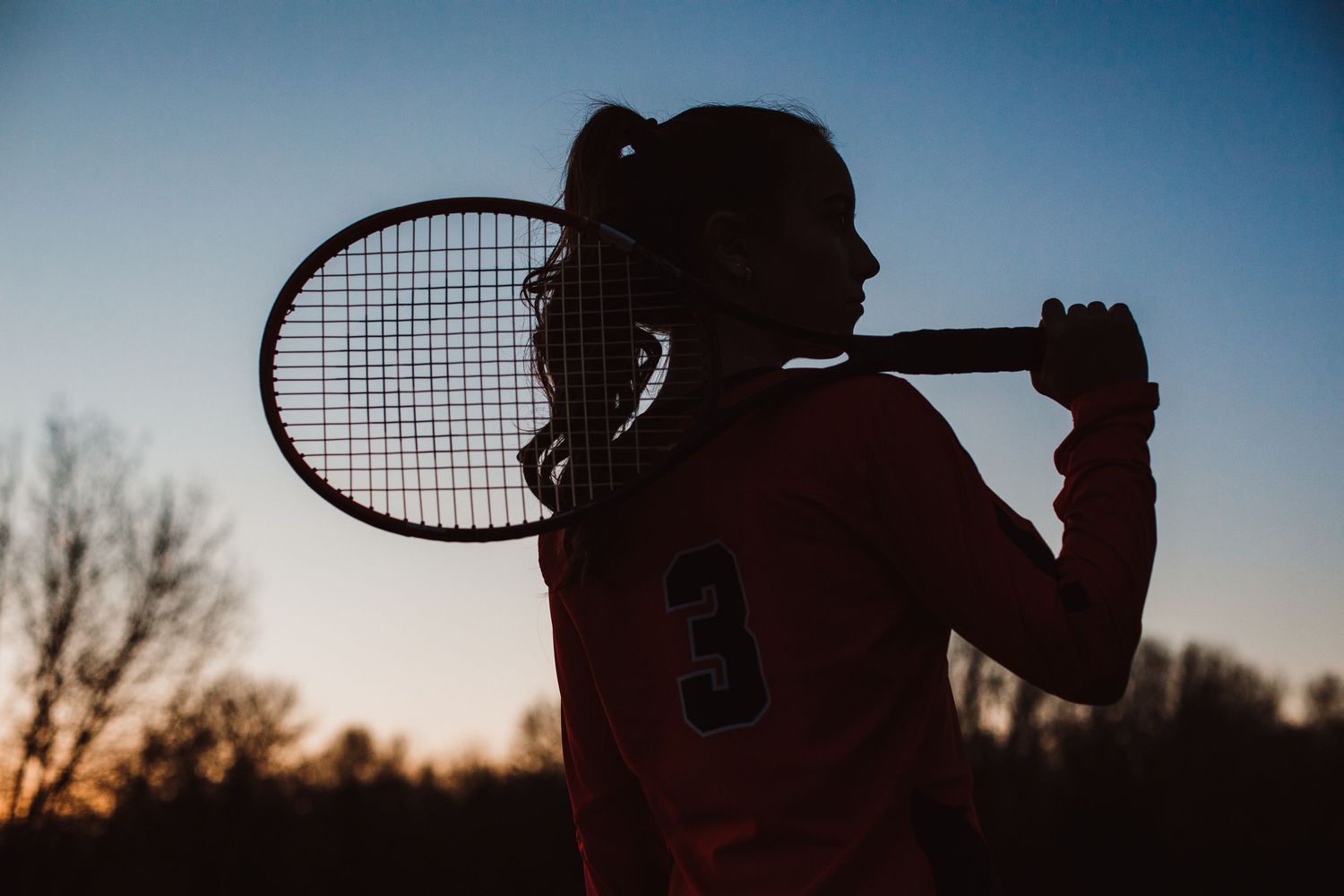 Silhouette of a young girl holding a tennis racket over her shoulder against a sunset sky.