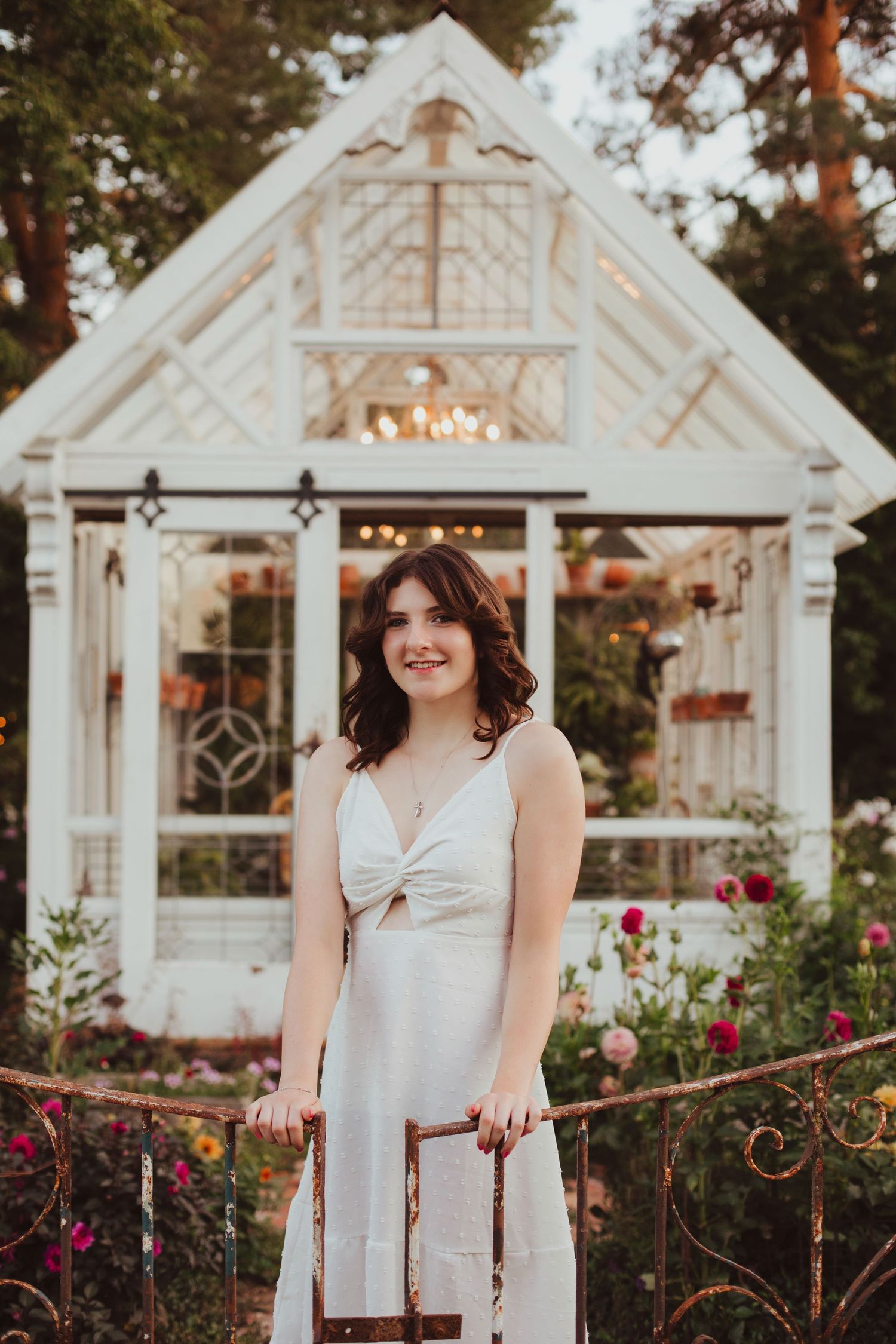 Young woman in white sundress standing by iron fence with a white glass greenhouse behind her.