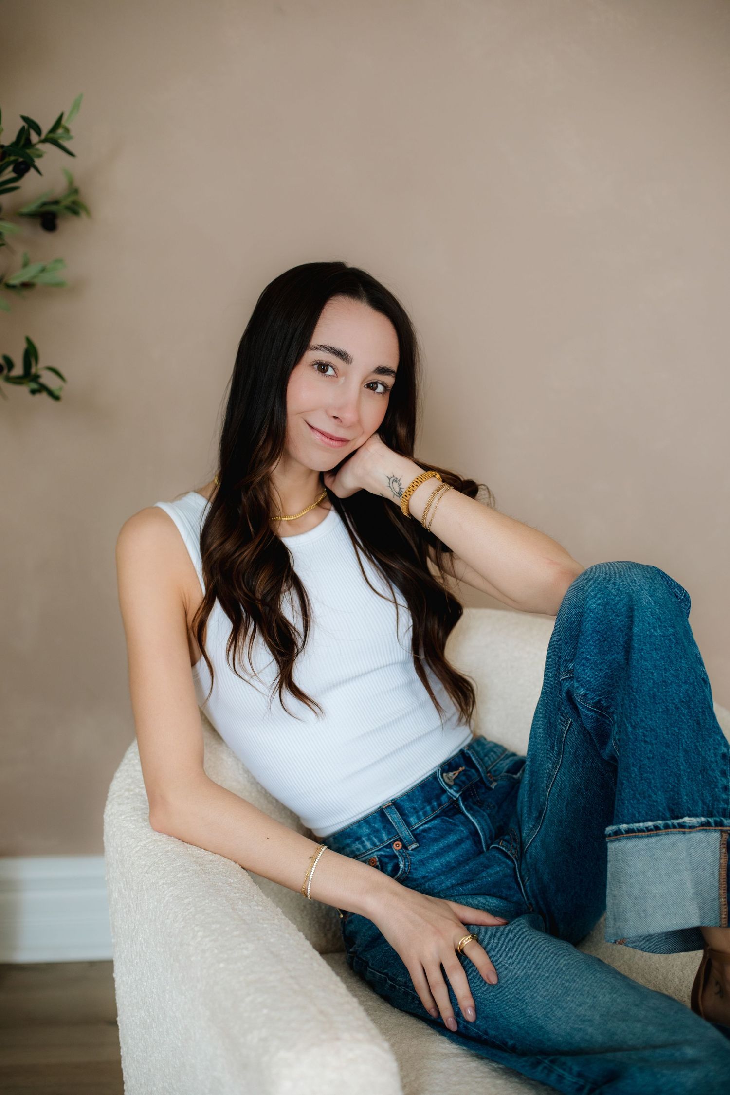 Brunette woman relaxing in white armchair wearing white tank top, jeans, and gold bracelets in neutral room.