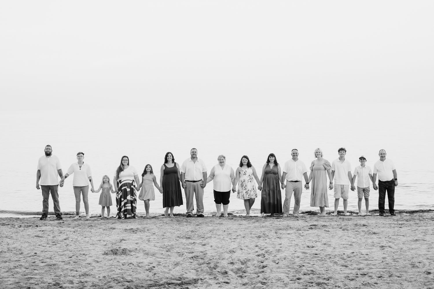 Black and white beach photo of a large multigenerational family holding hands in a long line by the ocean.