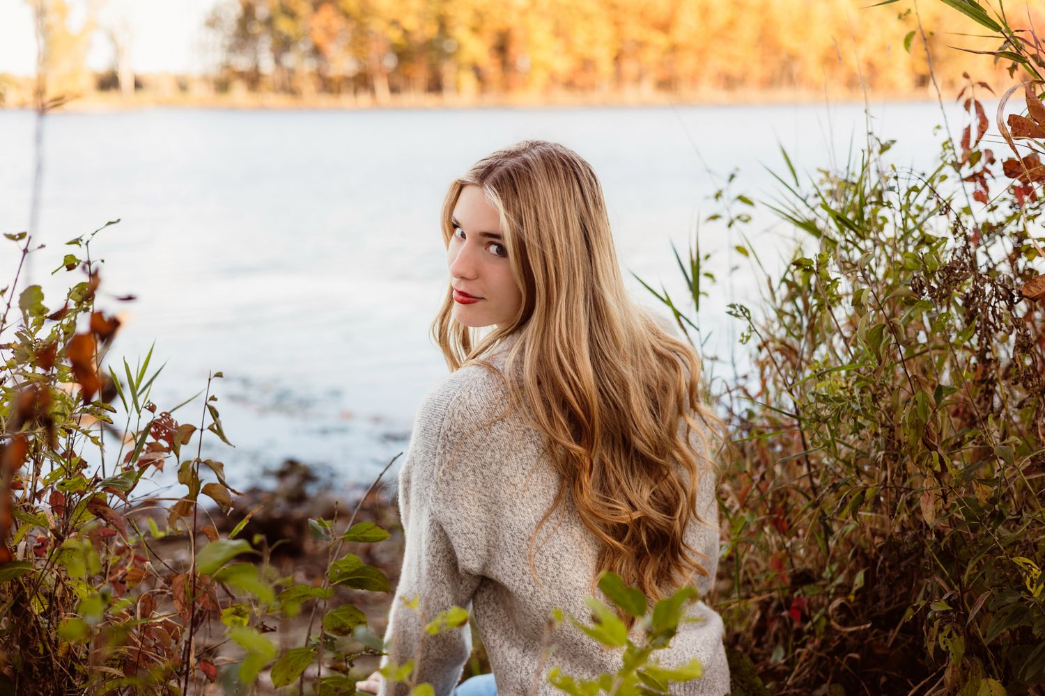 Young woman with long blonde hair in a cozy sweater sits by a lake surrounded by autumn foliage.