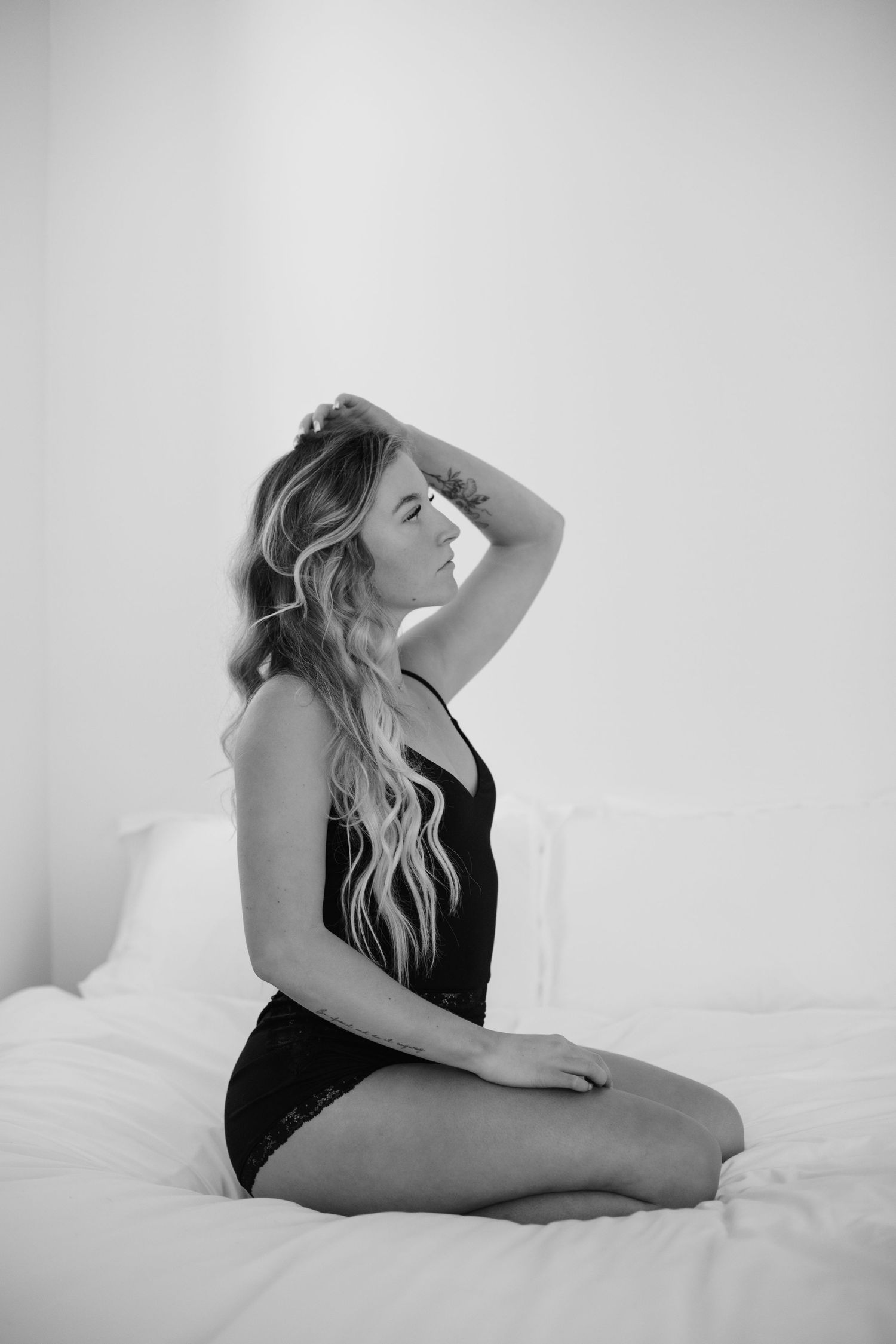 Black and white photo of blonde woman in black bodysuit sitting gracefully on white bed.