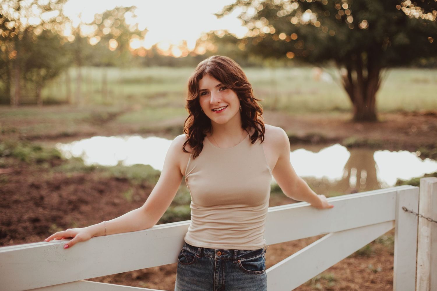 Young woman in beige tank top and jeans leaning on white fence with pond and trees in background.