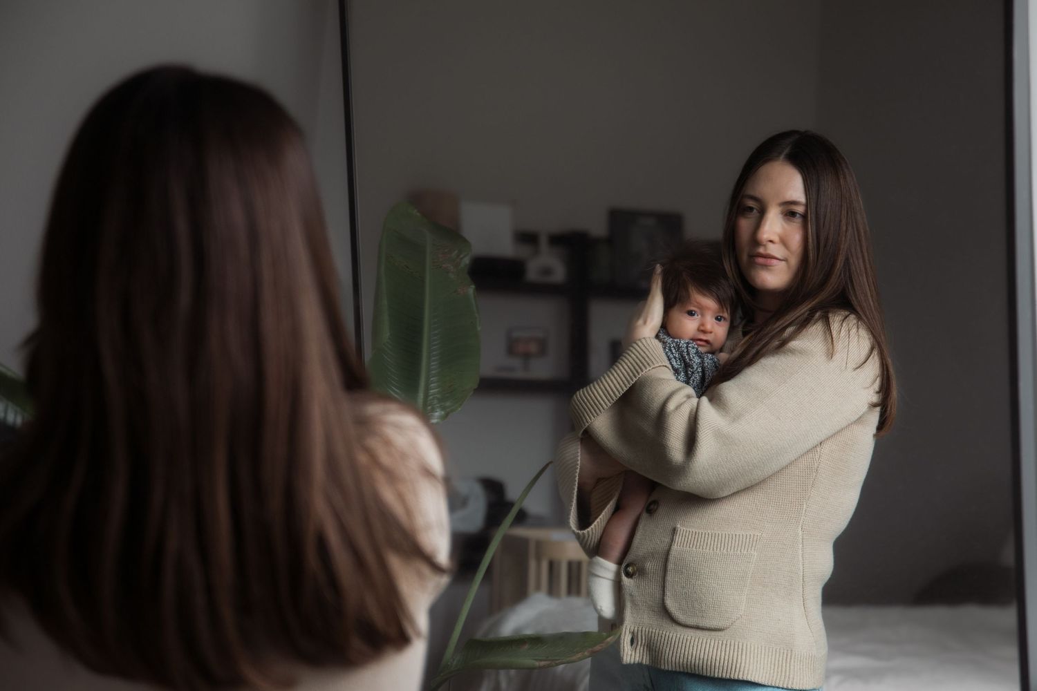 Mother holding sleeping baby while looking in mirror reflection at home.