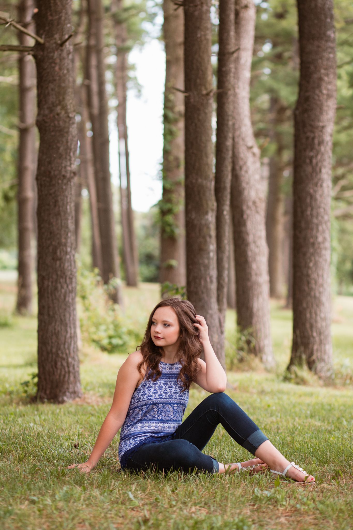 Young girl in blue top and jeans sitting on grass among tall pine trees in a serene outdoor setting.
