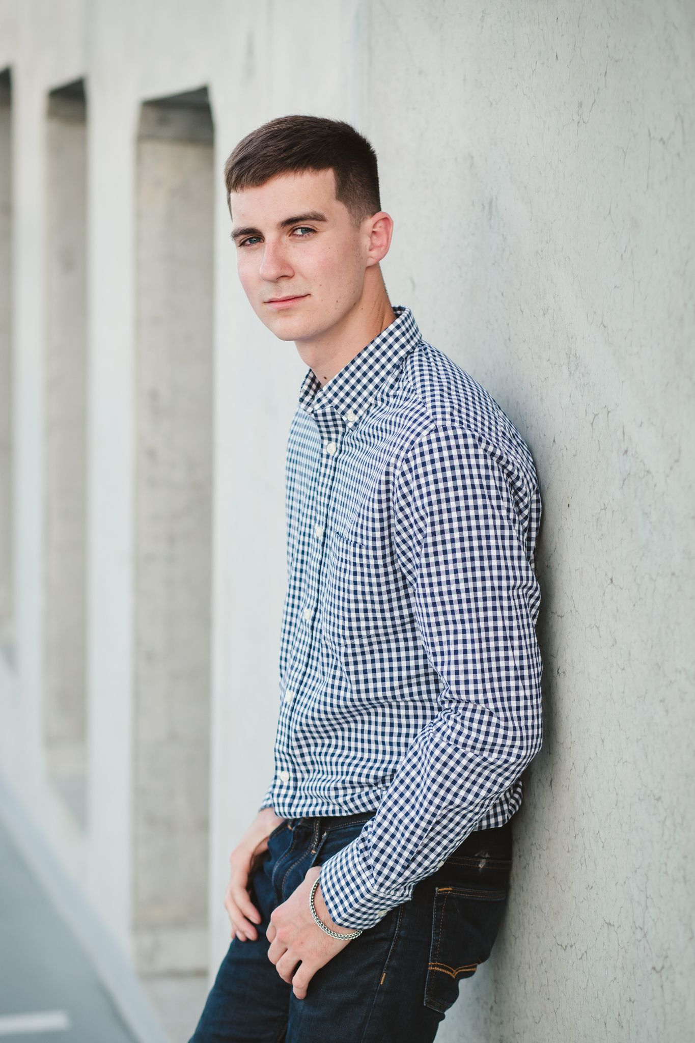Young man in checkered shirt and jeans leaning against a concrete wall outdoors, looking at camera.