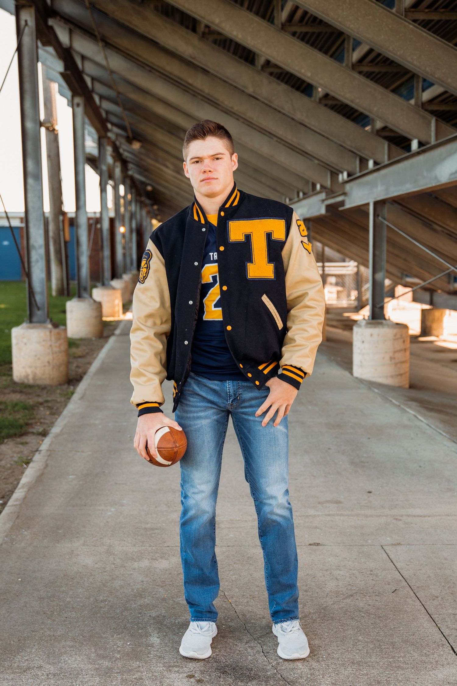 Young man in varsity letterman jacket holding football under stadium bleachers during senior portrait session.