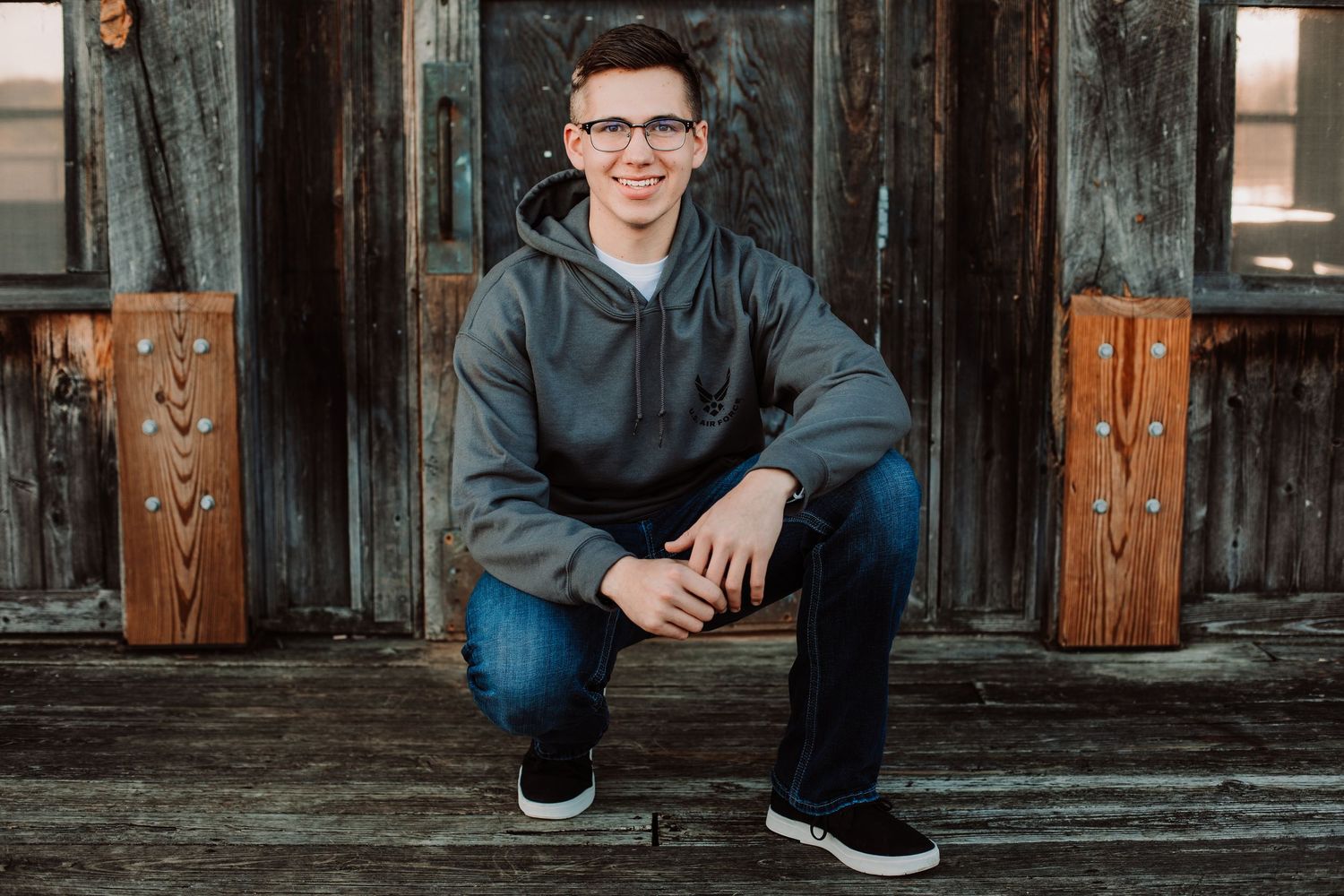 Young man in gray hoodie and jeans crouching on wooden steps, smiling, wearing glasses and black sneakers.
