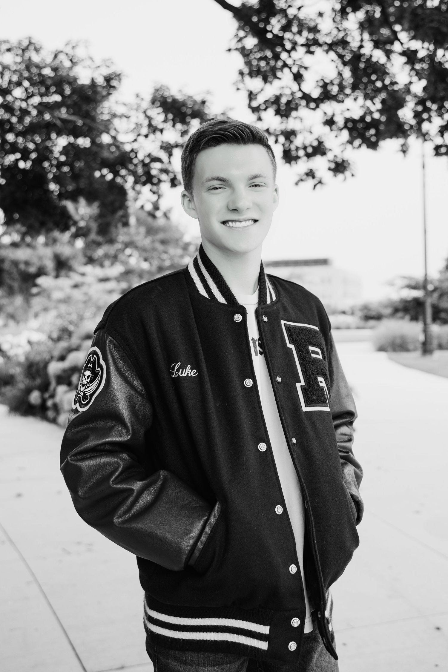 Smiling young man in a letterman jacket with leather sleeves posing outdoors in a black and white photo.