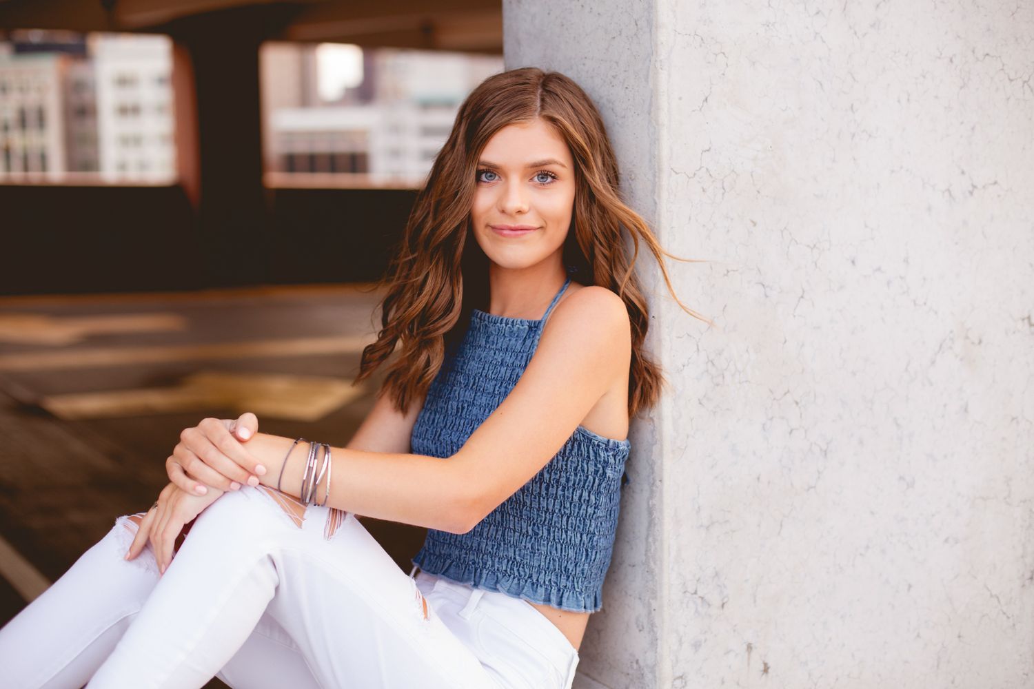Young woman in blue crop top and white jeans sits relaxed in urban parking structure.