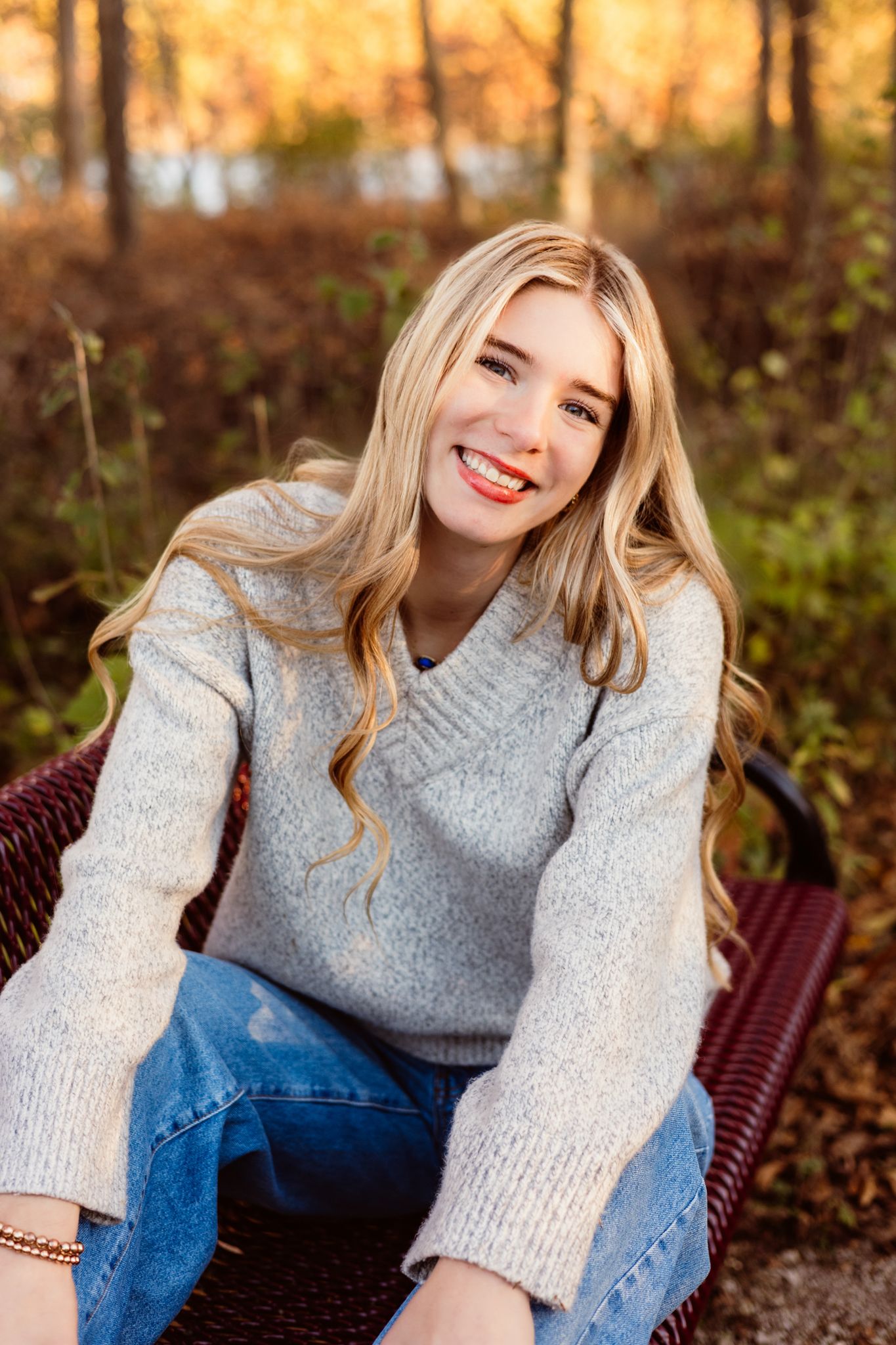 Smiling blonde woman in gray sweater and jeans sitting on a bench outdoors in autumn.