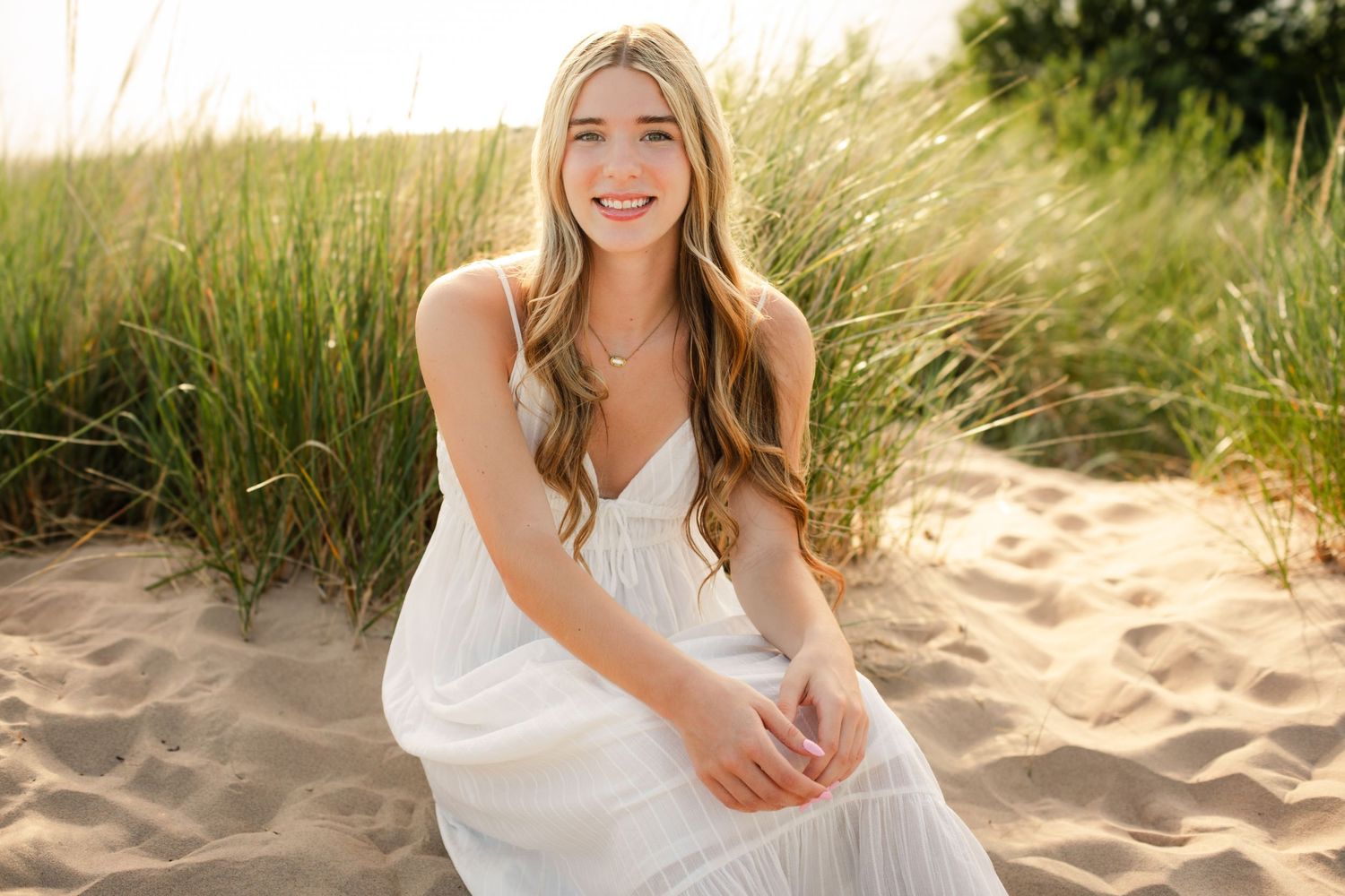 Smiling young woman in white dress sitting on sandy beach dunes with tall green grass in golden hour light.