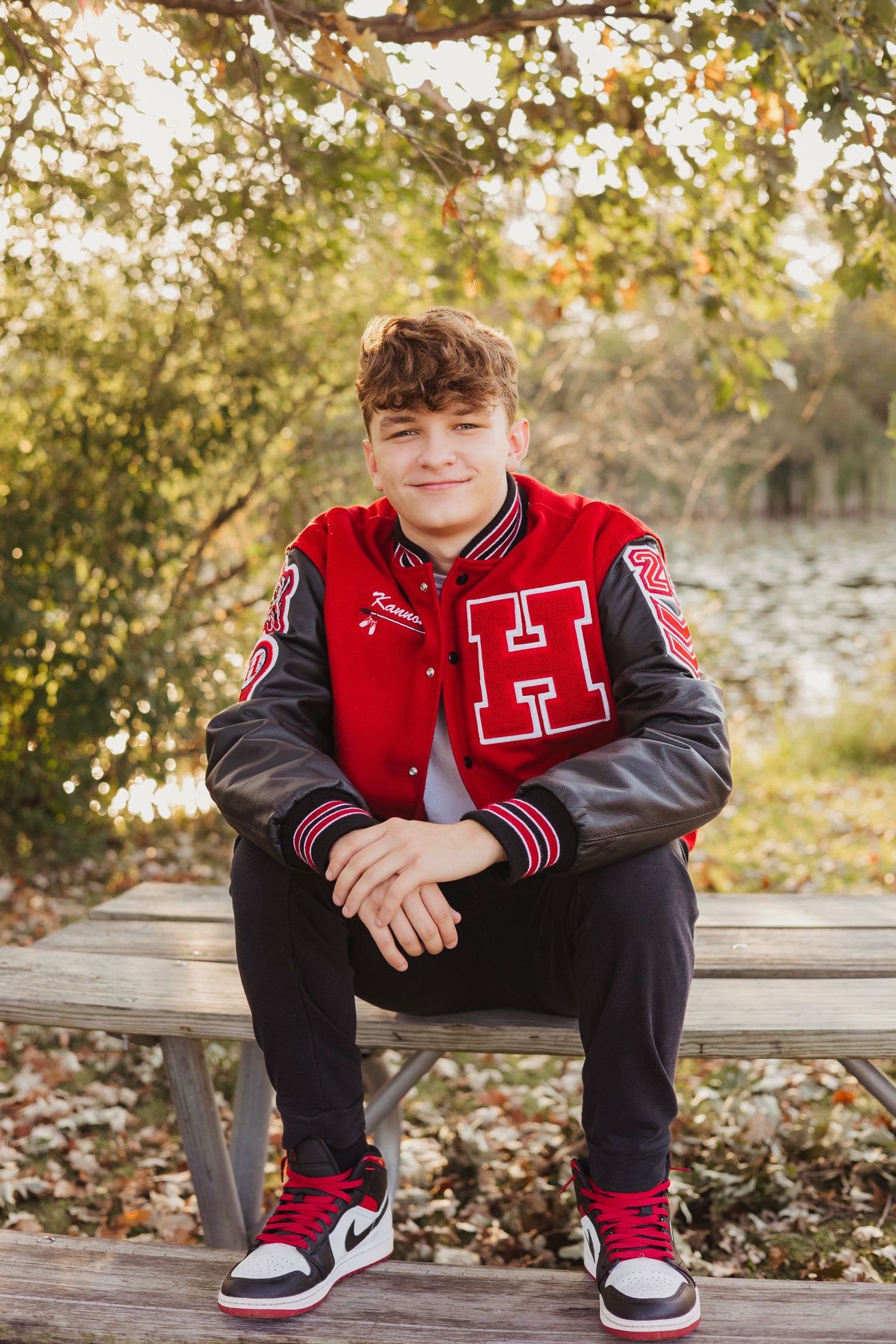 Young man in red varsity jacket with H letter sits on picnic bench outdoors among autumn foliage.