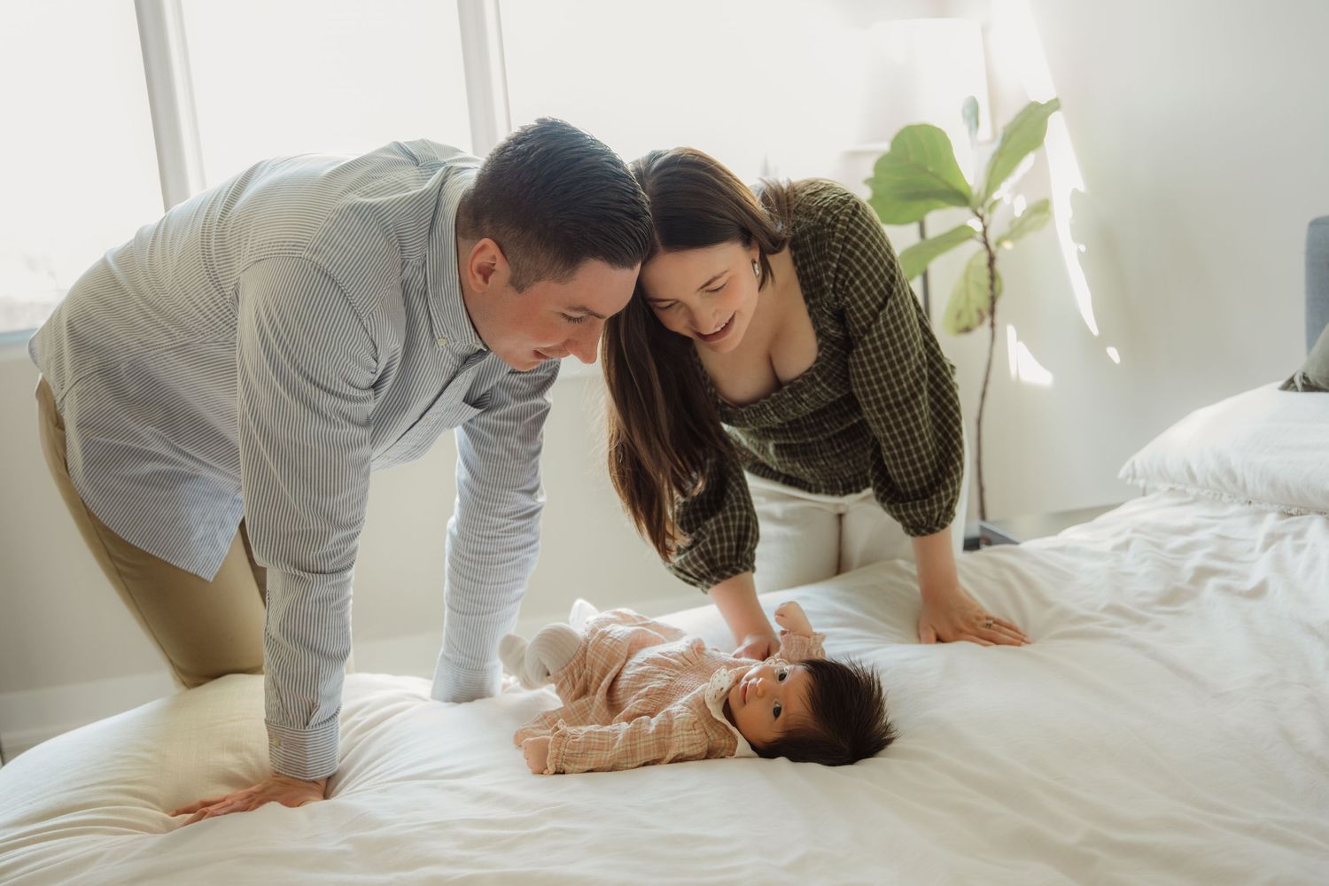 Happy parents leaning over newborn baby lying on white bed together.