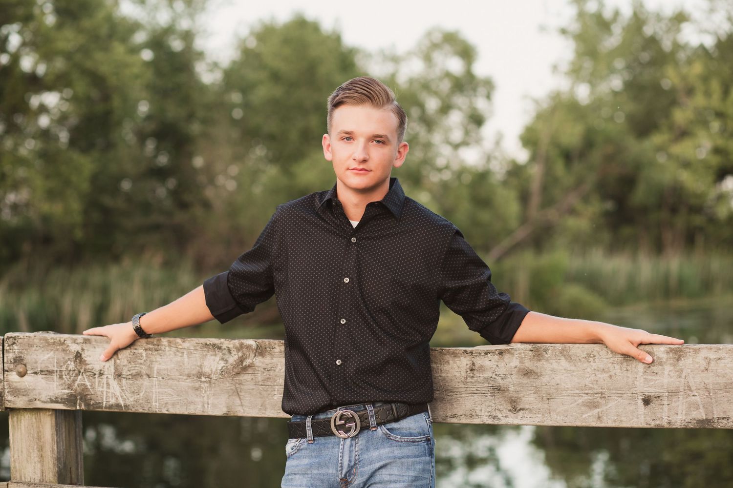 Young man in black shirt and jeans leaning on rustic wooden fence near pond with green trees background.