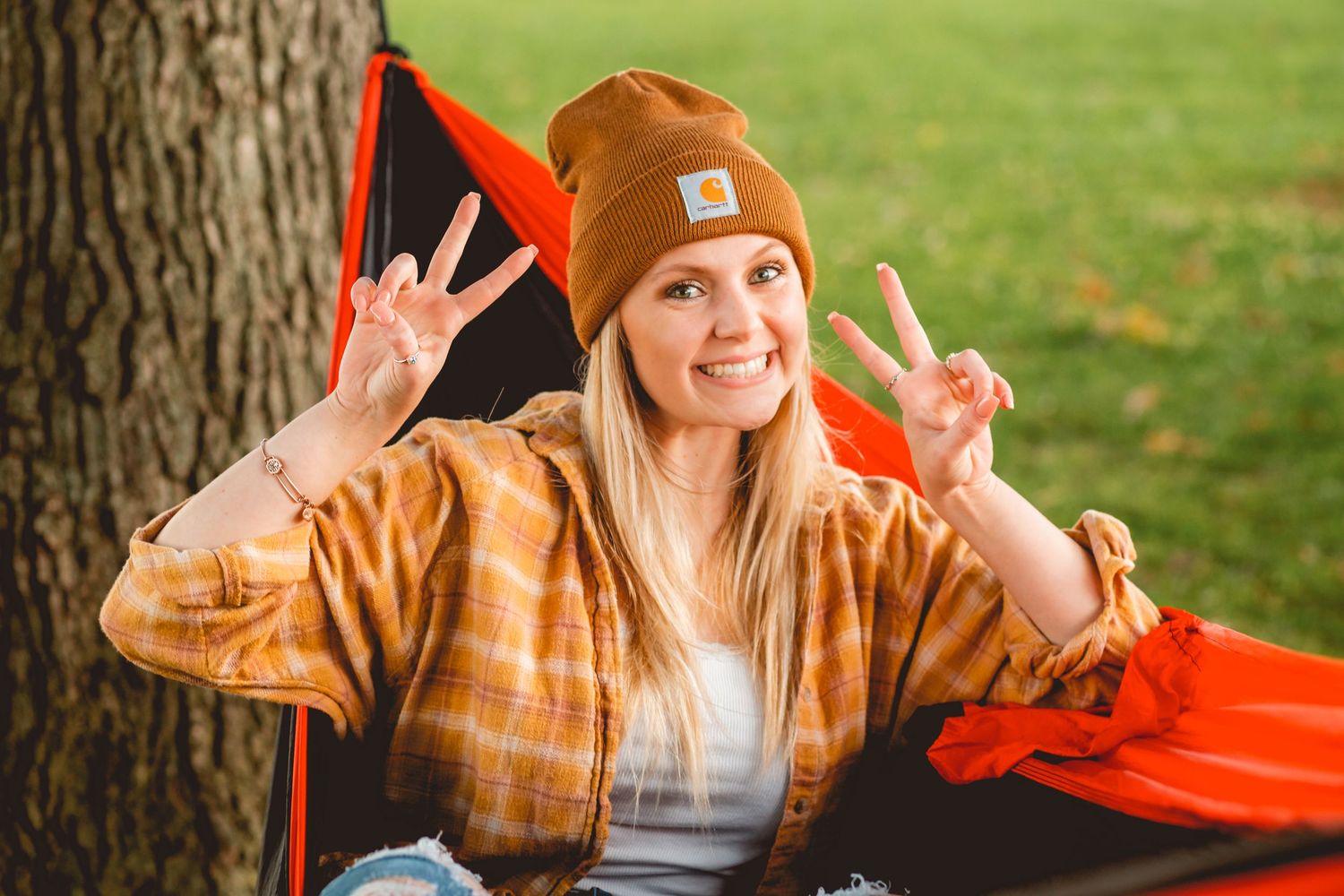 Cheerful blonde woman in plaid shirt and brown beanie making peace signs in hammock.